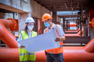 Two technicians in hard hats hold plans at an industrial facility.