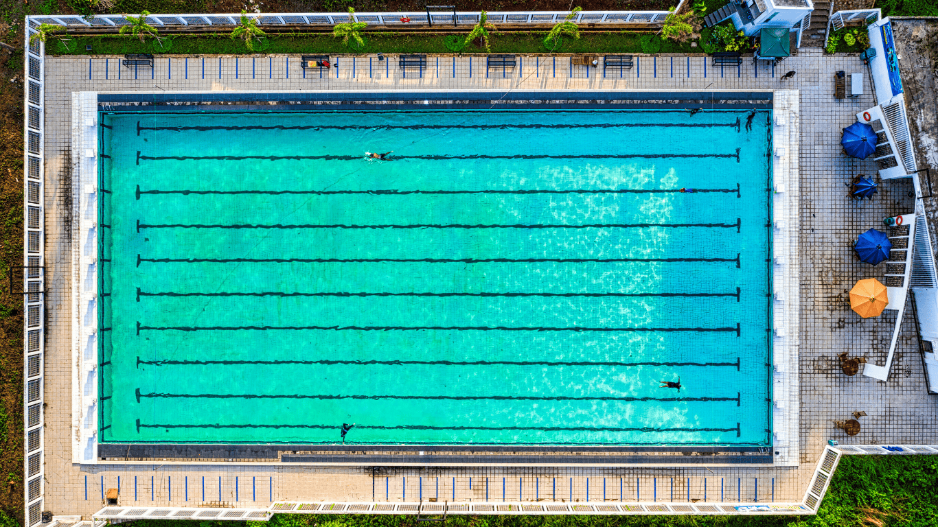 Aerial view of a large swimming pool at a hotel or resort, with clear turquoise water, swimming lanes, tiled walkways, and a surrounding pool deck.
